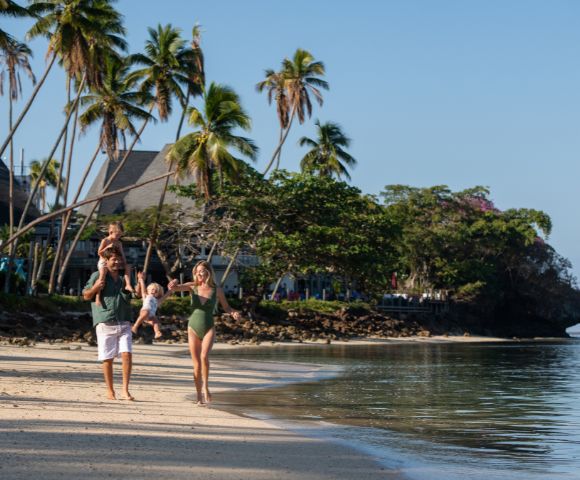 A joyful family walks along a tropical beach with palm trees. The father carries a child on his shoulders, while the mother holds hands with another child.