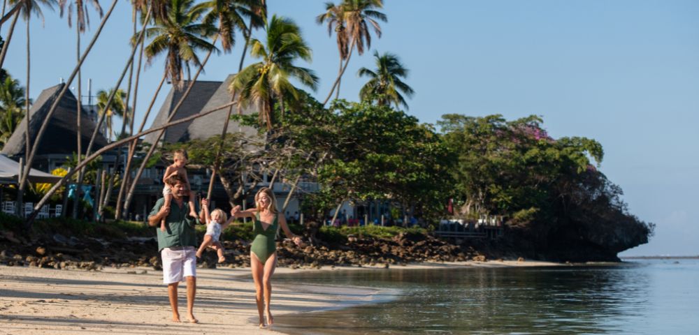 A joyful family walks along a tropical beach with palm trees. The father carries a child on his shoulders, while the mother holds hands with another child.