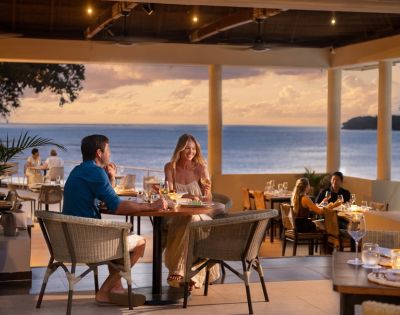 Couple dining at an oceanfront restaurant during sunset. They are smiling, enjoying a romantic ambiance with soft lighting and an ocean view.