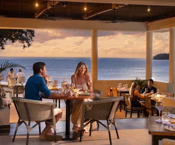 Couple dining at an oceanfront restaurant during sunset. They are smiling, enjoying a romantic ambiance with soft lighting and an ocean view.
