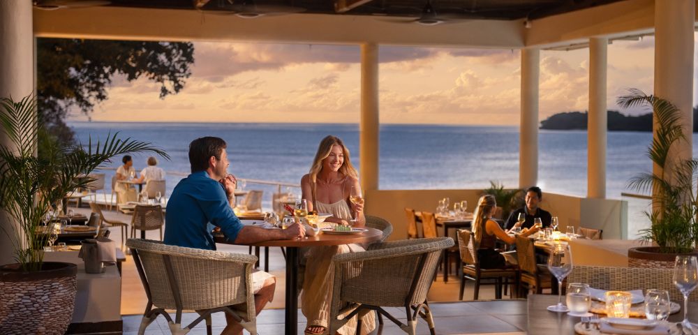 Couple dining at an oceanfront restaurant during sunset. They are smiling, enjoying a romantic ambiance with soft lighting and an ocean view.