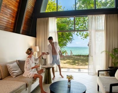 A couple enjoys a tropical retreat in a sunlit villa with beach view. The woman sits on a sofa, while the man pours drinks by open glass doors. Relaxed ambiance.