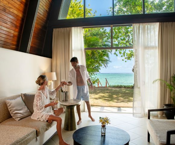 A couple enjoys a tropical retreat in a sunlit villa with beach view. The woman sits on a sofa, while the man pours drinks by open glass doors. Relaxed ambiance.