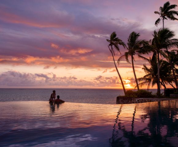 A couple stands in a serene infinity pool at sunset. The sky is painted with pink and purple hues, and palm trees are silhouetted against the horizon.