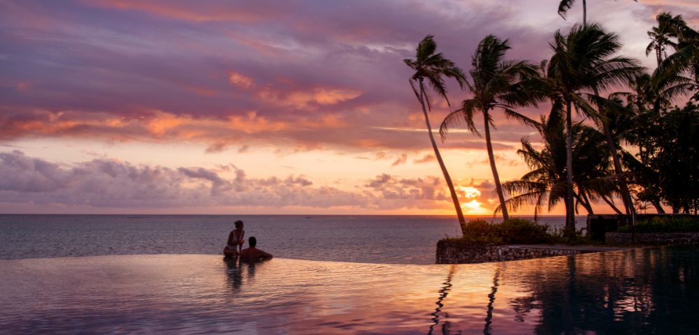 A couple stands in a serene infinity pool at sunset. The sky is painted with pink and purple hues, and palm trees are silhouetted against the horizon.