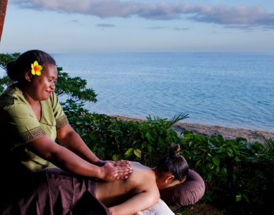 Woman receiving a relaxing back massage beside a tranquil ocean. The masseuse has a flower in her hair, enhancing the serene, tropical setting.