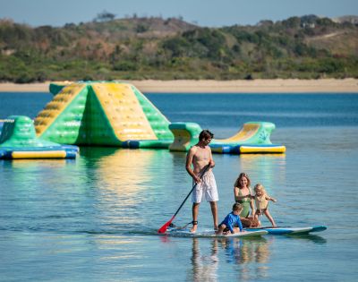 A man paddleboards with three children on a calm lake. Behind them, a colorful inflatable water park floats. The scene is joyful and serene.