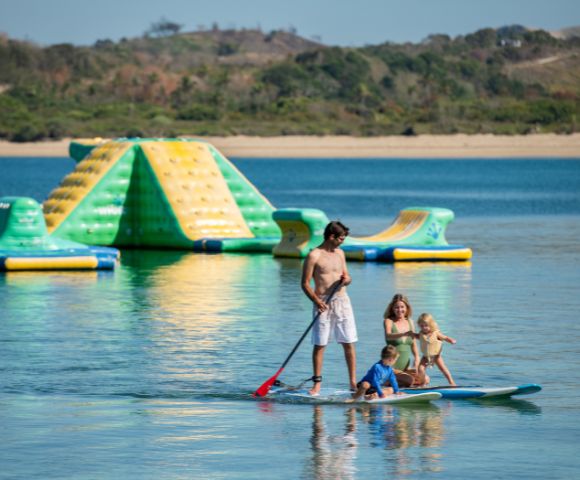 A man paddleboards with three children on a calm lake. Behind them, a colorful inflatable water park floats. The scene is joyful and serene.