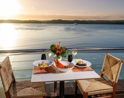 Outdoor table set with a seafood meal, wine glasses, and floral centerpiece overlooking a serene lake at sunset, evoking a romantic and tranquil ambiance.