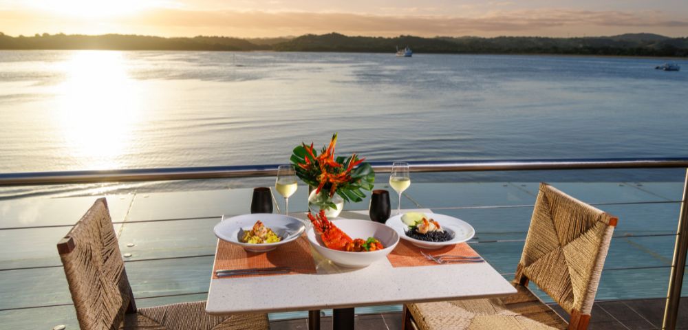 Outdoor table set with a seafood meal, wine glasses, and floral centerpiece overlooking a serene lake at sunset, evoking a romantic and tranquil ambiance.