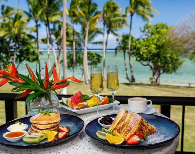 Breakfast on a sunny balcony overlooking a tropical beach. Plates with pancakes, French toast, fruits; champagne flutes and a bird of paradise flower.