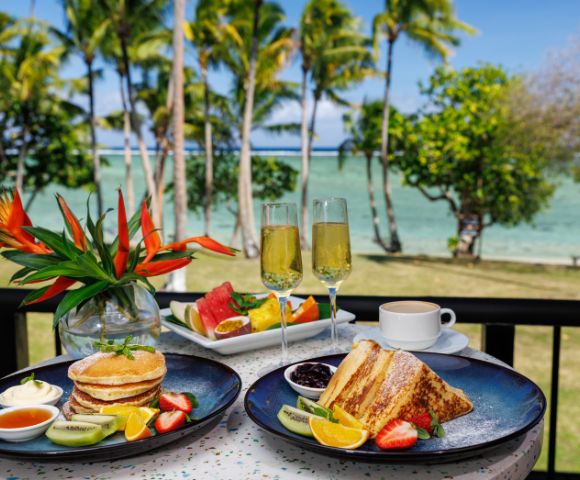 Breakfast on a sunny balcony overlooking a tropical beach. Plates with pancakes, French toast, fruits; champagne flutes and a bird of paradise flower.