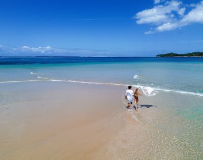 A couple walks hand in hand along a serene, sandy beach with clear turquoise waters under a vibrant blue sky, conveying tranquility and romance.