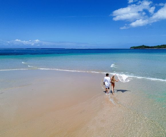 A couple walks hand in hand along a serene, sandy beach with clear turquoise waters under a vibrant blue sky, conveying tranquility and romance.