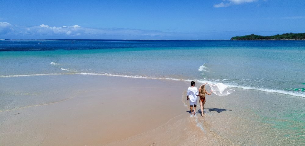 A couple walks hand in hand along a serene, sandy beach with clear turquoise waters under a vibrant blue sky, conveying tranquility and romance.