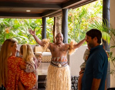 Dancer in traditional attire with grass skirt and arm bands joyfully welcomes a family at a tropical resort entrance, surrounded by lush greenery.