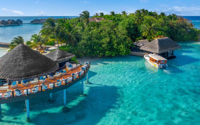 Overwater bungalows surrounded by turquoise waters, with a boat near a tropical island and lush greenery under a sunny sky.