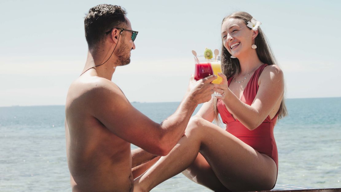 A couple smiling and toasting with cocktails by the ocean, surrounded by tropical scenery. The mood is joyful and relaxed, perfect for a vacation.