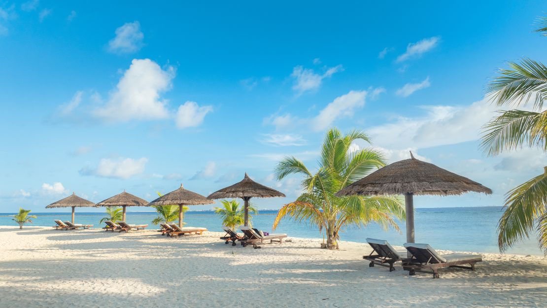 Tropical beach scene with thatched umbrellas and lounge chairs on white sand, surrounded by palm trees. A calm ocean under a clear blue sky suggests relaxation.