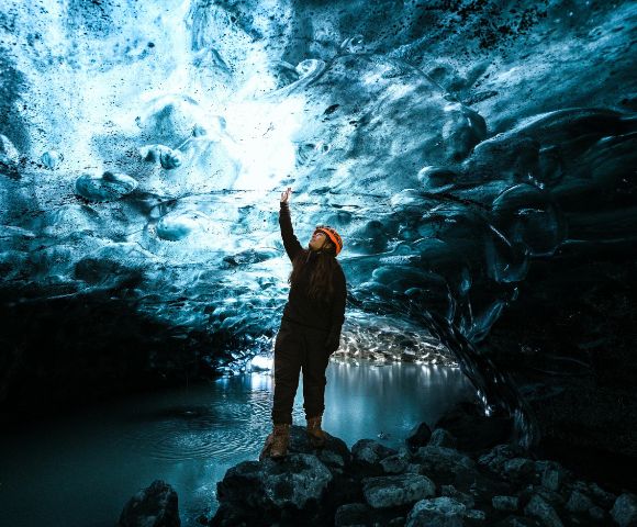 Person stands inside an ice cave, reaching up to the textured, illuminated blue ice ceiling. Reflective water and rocks create a serene, wondrous atmosphere.