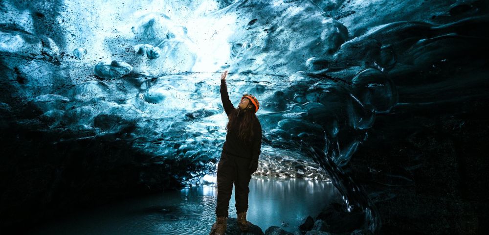 Person stands inside an ice cave, reaching up to the textured, illuminated blue ice ceiling. Reflective water and rocks create a serene, wondrous atmosphere.