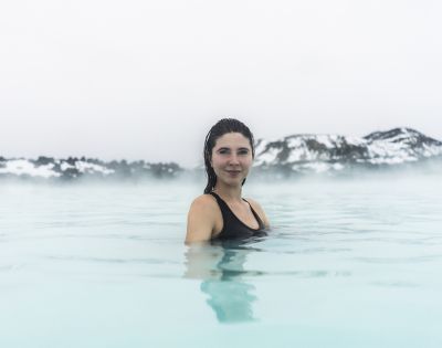 A woman in a black swimsuit smiles while standing in a steaming, pale blue geothermal pool, set against a foggy backdrop of snow-covered hills.