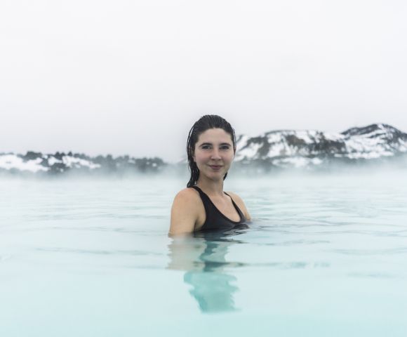 A woman in a black swimsuit smiles while standing in a steaming, pale blue geothermal pool, set against a foggy backdrop of snow-covered hills.