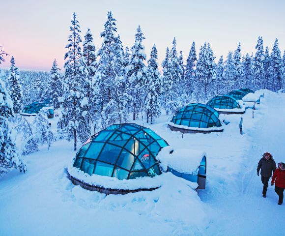 Snow-covered glass igloos nestled in a wintery forest. Two people walk nearby under a pastel sky, conveying tranquility and wonder.