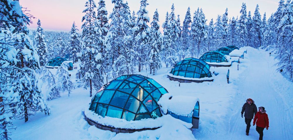Snow-covered glass igloos nestled in a wintery forest. Two people walk nearby under a pastel sky, conveying tranquility and wonder.