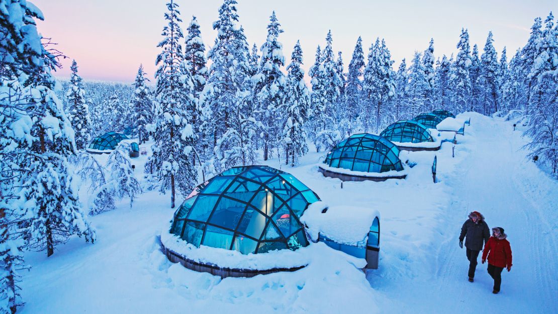 Snow-covered glass igloos nestled in a wintery forest. Two people walk nearby under a pastel sky, conveying tranquility and wonder.