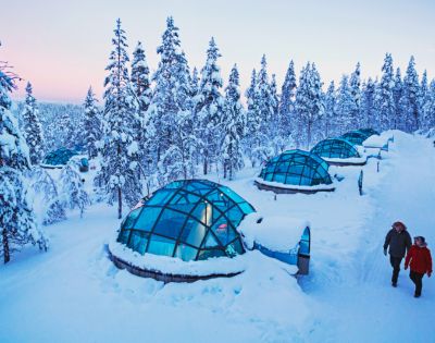Snow-covered glass igloos nestled in a wintery forest. Two people walk nearby under a pastel sky, conveying tranquility and wonder.