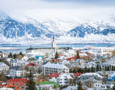 Snow-covered town under a bright blue sky, with colorful rooftops scattered among buildings. Majestic snow-capped mountains rise in the background, conveying tranquility.