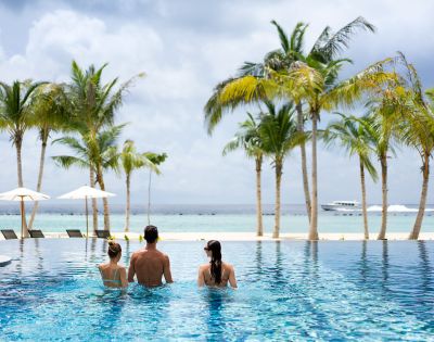 Three people in swimsuits relax on the edge of an infinity pool facing a tranquil ocean. Palm trees and a boat enhance the tropical vibe.
