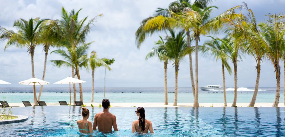 Three people in swimsuits relax on the edge of an infinity pool facing a tranquil ocean. Palm trees and a boat enhance the tropical vibe.