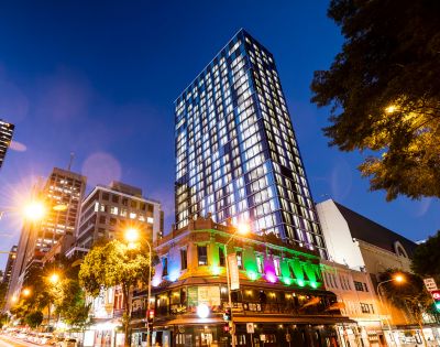 A tall, modern skyscraper stands against a twilight sky, illuminated with bright windows. Below, a historic building glows with vibrant green and purple lights.