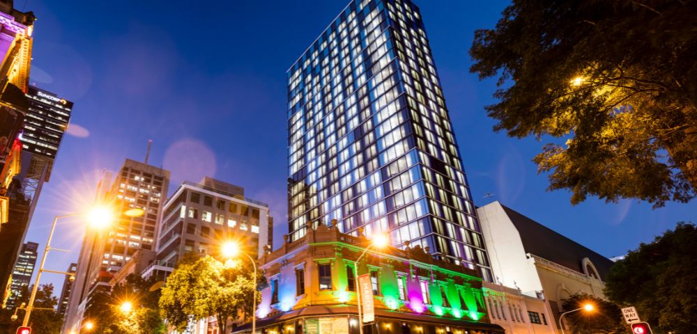 A tall, modern skyscraper stands against a twilight sky, illuminated with bright windows. Below, a historic building glows with vibrant green and purple lights.