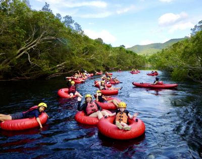 A group of people wearing helmets and life vests float down a scenic river on red inner tubes, surrounded by lush greenery and hills under a bright sky.