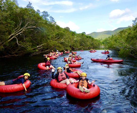 A group of people wearing helmets and life vests float down a scenic river on red inner tubes, surrounded by lush greenery and hills under a bright sky.