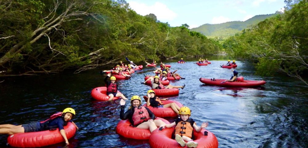 A group of people wearing helmets and life vests float down a scenic river on red inner tubes, surrounded by lush greenery and hills under a bright sky.