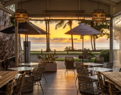 Outdoor cafe with empty tables and chairs facing a serene ocean sunset. Palm trees silhouette against an orange sky, creating a peaceful atmosphere.