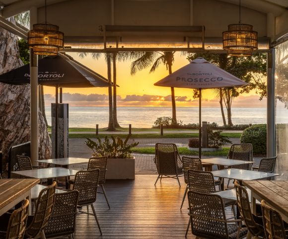 Outdoor cafe with empty tables and chairs facing a serene ocean sunset. Palm trees silhouette against an orange sky, creating a peaceful atmosphere.