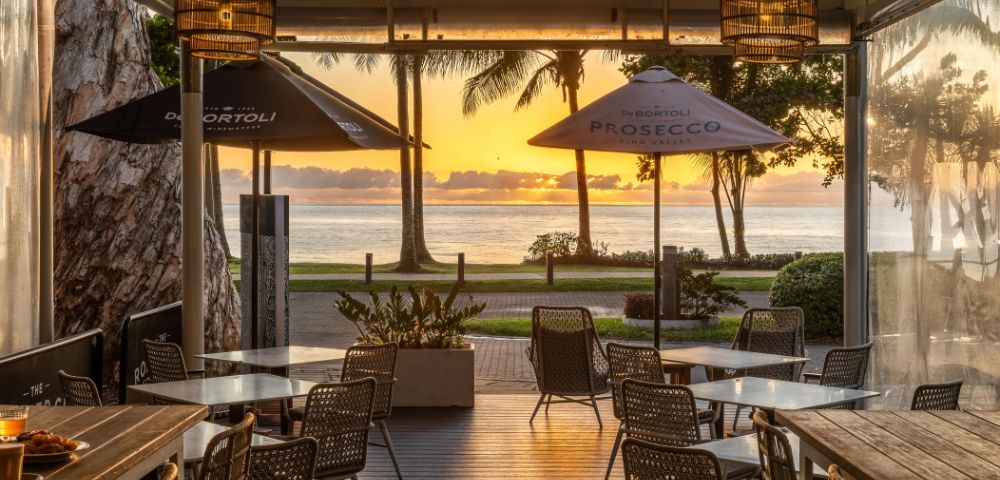 Outdoor cafe with empty tables and chairs facing a serene ocean sunset. Palm trees silhouette against an orange sky, creating a peaceful atmosphere.