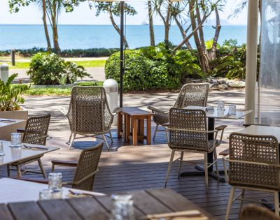 Outdoor cafe with wicker chairs and tables set for dining, overlooking a scenic ocean view. The atmosphere is calm and inviting, with lush greenery.