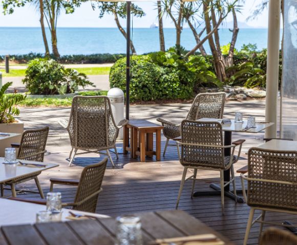 Outdoor cafe with wicker chairs and tables set for dining, overlooking a scenic ocean view. The atmosphere is calm and inviting, with lush greenery.