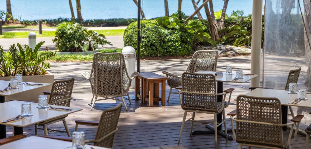 Outdoor cafe with wicker chairs and tables set for dining, overlooking a scenic ocean view. The atmosphere is calm and inviting, with lush greenery.