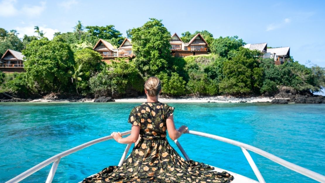 Woman in floral dress sits on boat, facing lush island with wooden houses. Clear turquoise water reflects a sunny, tranquil atmosphere.