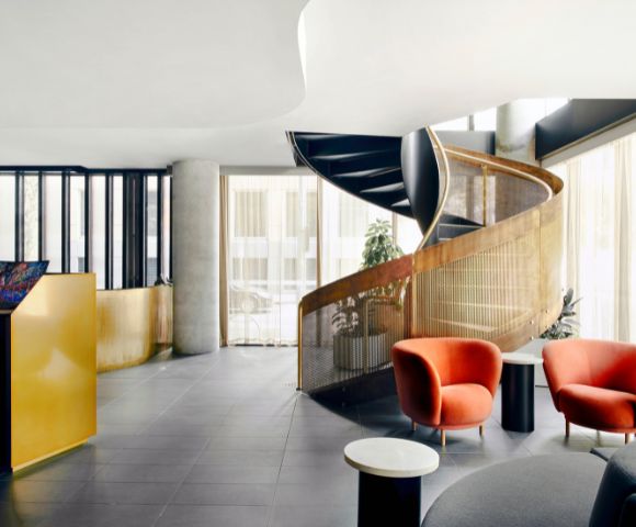 Modern interior with a sweeping spiral staircase, gold railing, and natural light. Features two vibrant red chairs and sleek black tables.