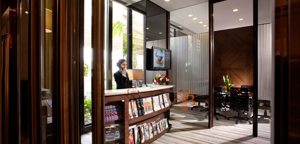 Modern office reception with a smiling person at the front desk, magazines displayed, glass walls, and a meeting room with a table and chairs in the background.