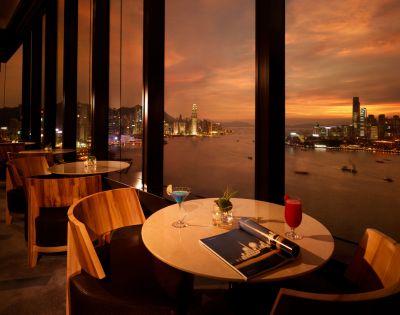 Upscale restaurant with large windows overlooks a harbor at sunset. Two tables have drinks, a magazine, and elegant décor, creating a warm, sophisticated ambiance.