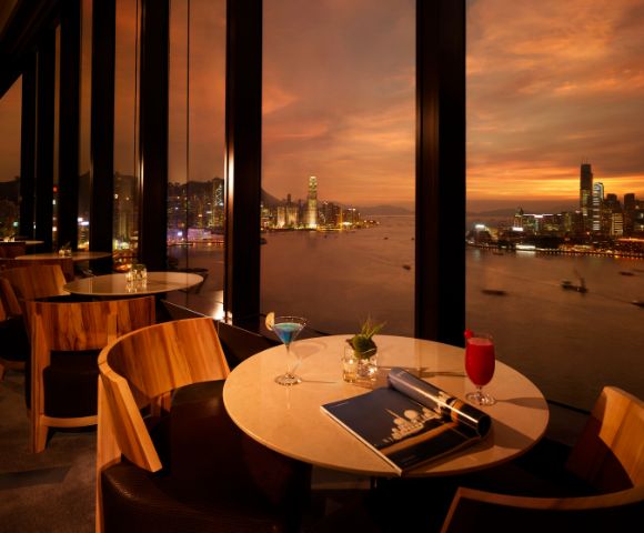 Upscale restaurant with large windows overlooks a harbor at sunset. Two tables have drinks, a magazine, and elegant décor, creating a warm, sophisticated ambiance.